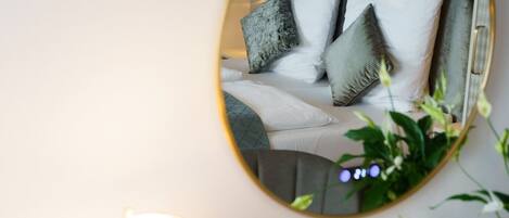 A bedside table with a lamp, a small vase, and a mirror reflecting a bed with pillows.