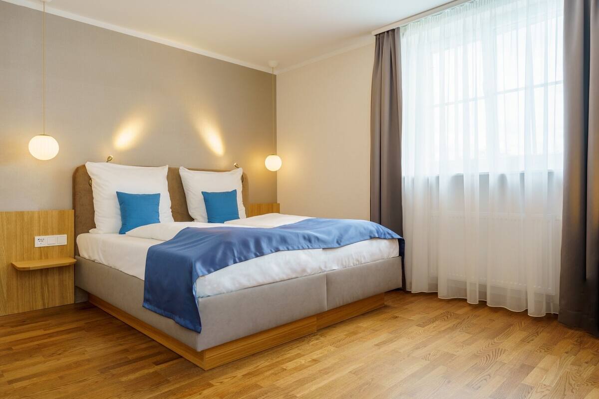 Un dormitorio moderno con piso de madera, una cama con ropa de cama blanca y azul, dos lámparas de noche y un ventanal con cortinas translúcidas.