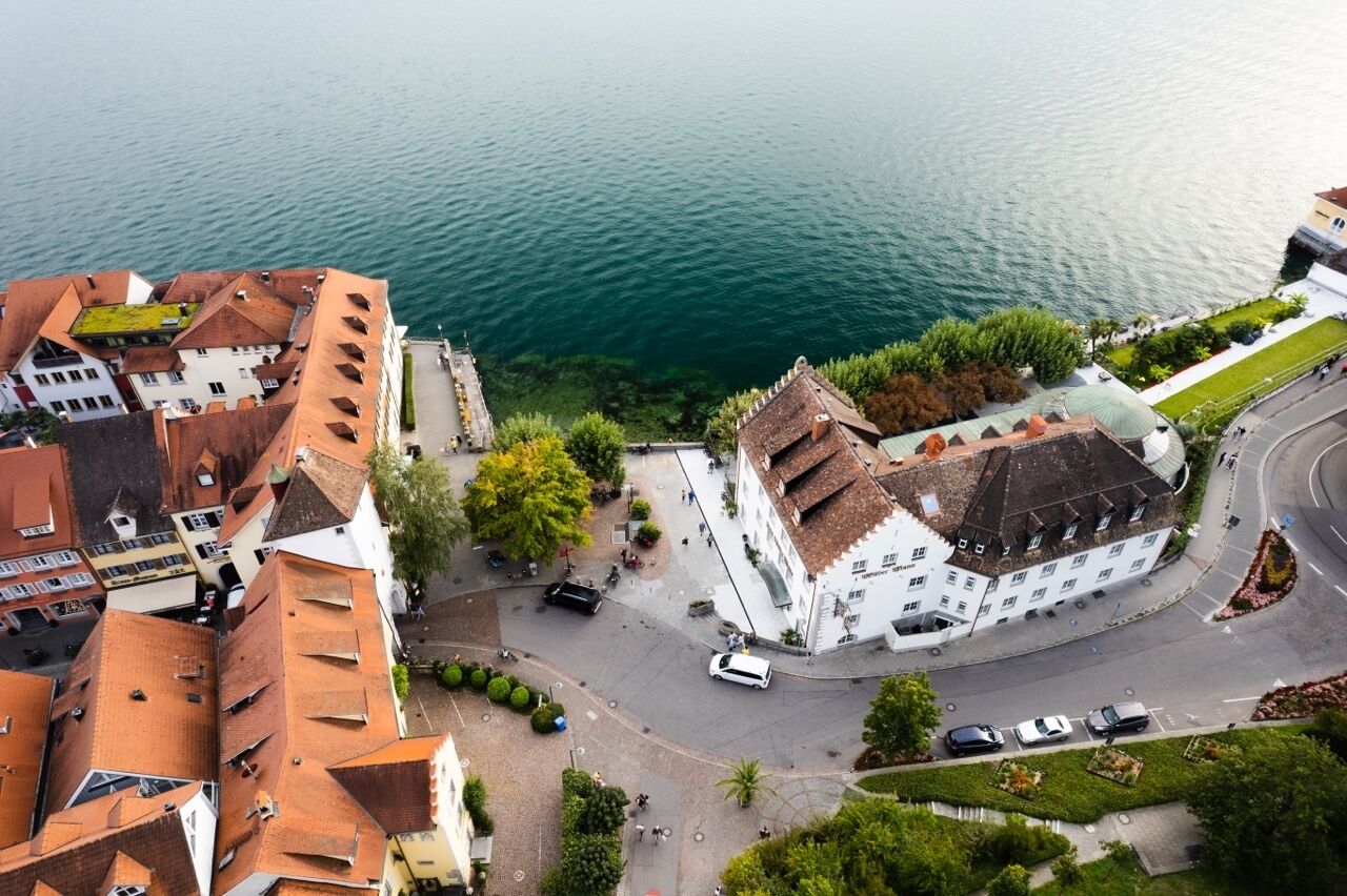 Vista aérea de un pueblo histórico con un edificio blanco destacado y techo verde, rodeado de otros edificios y una masa de agua.
