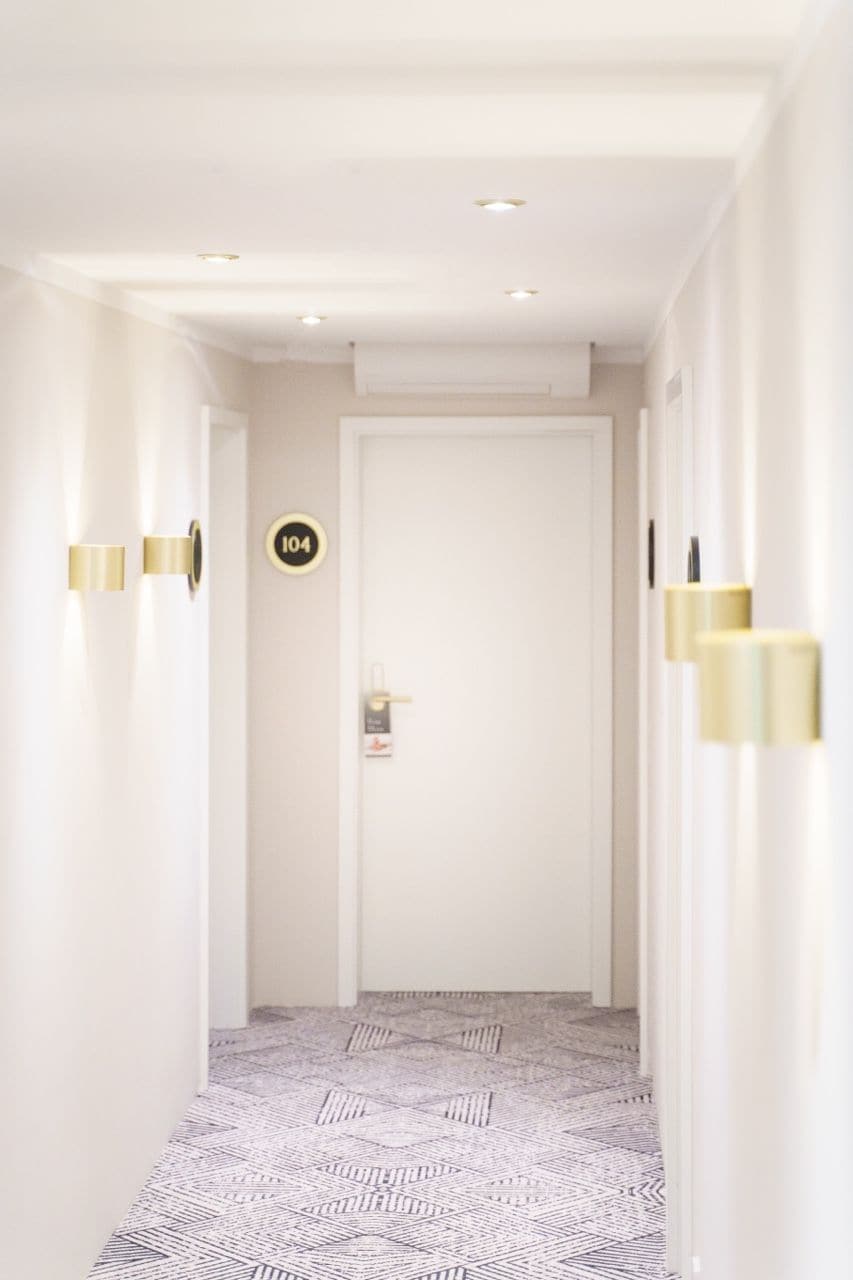Un couloir doté d’un sol à motifs, une porte avec une plaque numérotée et des luminaires fixés au mur.