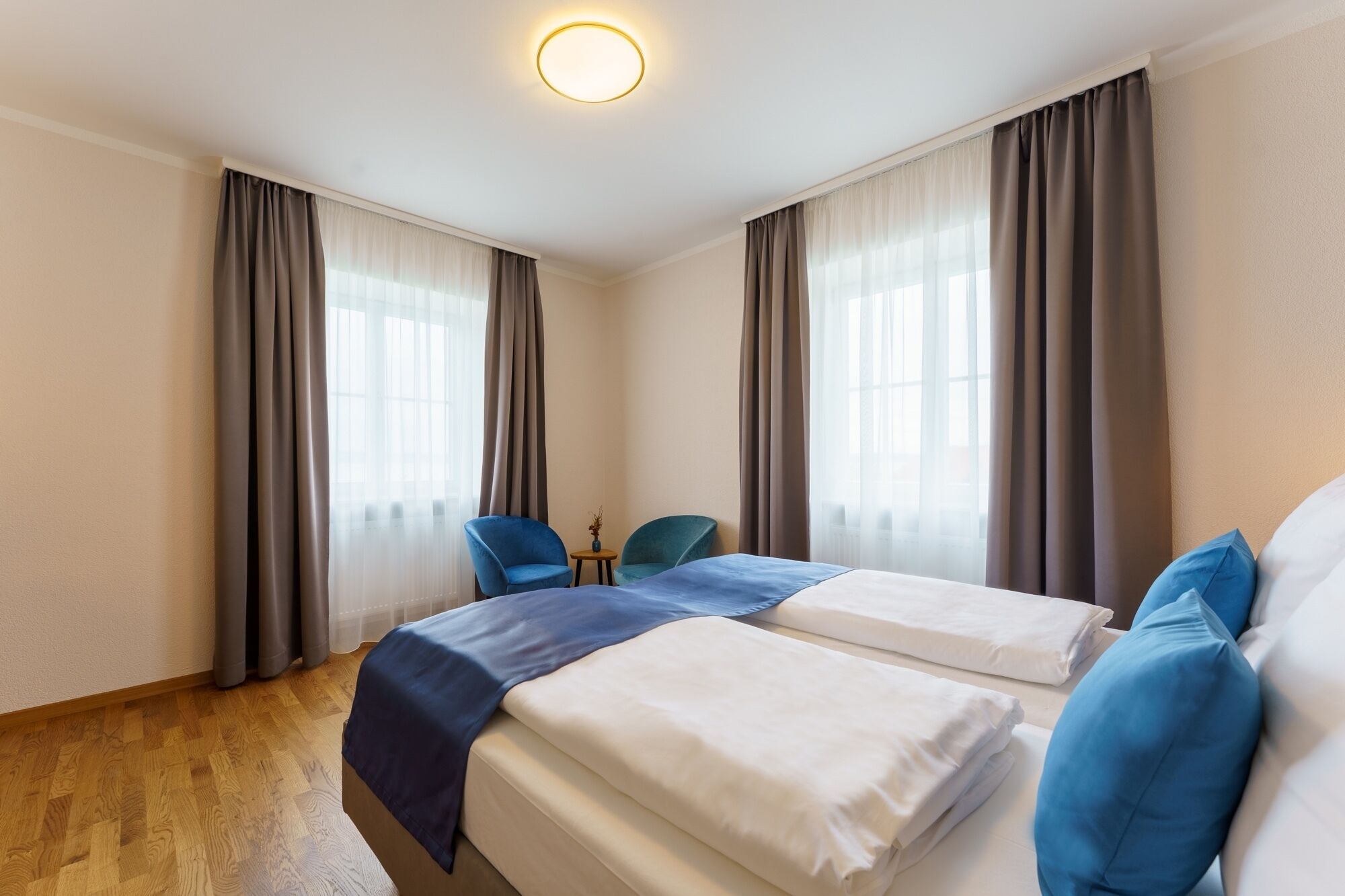 Quarto de hotel com duas camas, uma mesa pequena, duas cadeiras e janelas amplas com cortinas.
