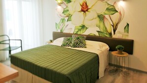A bedroom with a large floral mural on the wall, a green bedspread, a small round table with a plant, and a hanging light fixture.