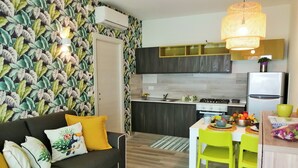 A modern kitchen with a dining area, featuring a grey sofa with yellow cushions, a bright yellow dining table, and a hanging light fixture.