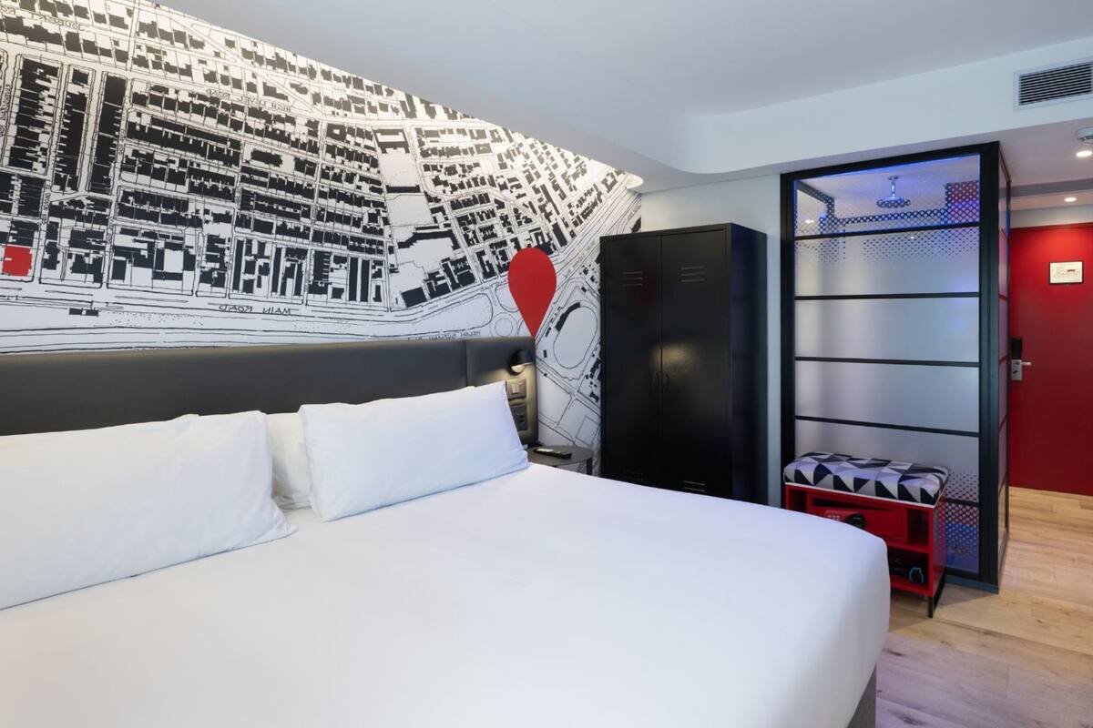 A hotel room with a large bed, a map mural on the wall, a red door, and a seating area with a red bench.