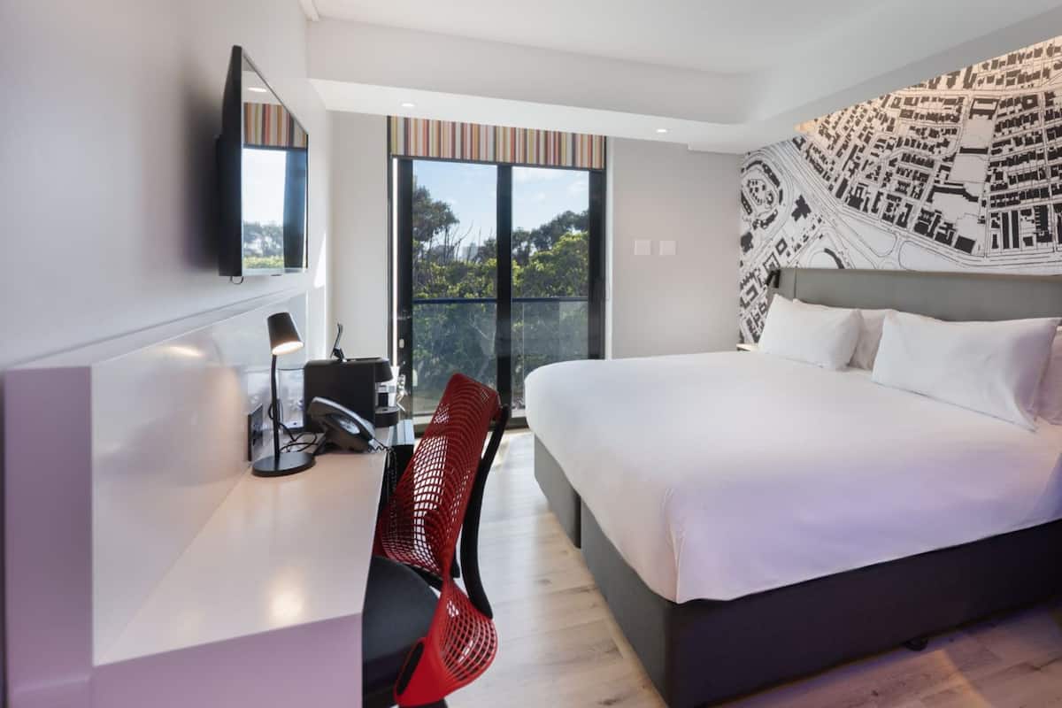 A modern hotel room with a large bed, a desk with a chair, a TV mounted on the wall, and a cityscape mural on the wall.
