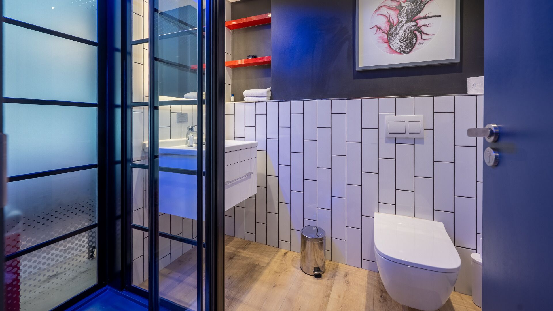 Un baño moderno con entrada de puerta de vidrio, paredes de azulejos blancos, suelo de madera, inodoro blanco y lavamanos con espejo.