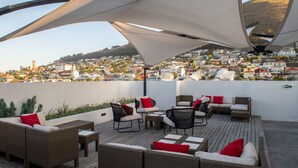 A rooftop terrace with wicker furniture, red cushions, and a view of a hillside town. - ANEW Hotel Green Point (Cape Town)