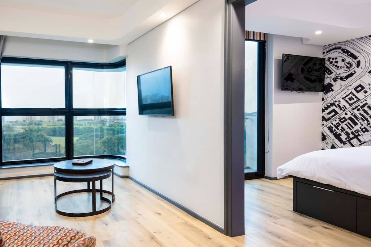 A modern hotel room with a large window, a flat-screen TV, a bed, a small round table, and a potted plant.