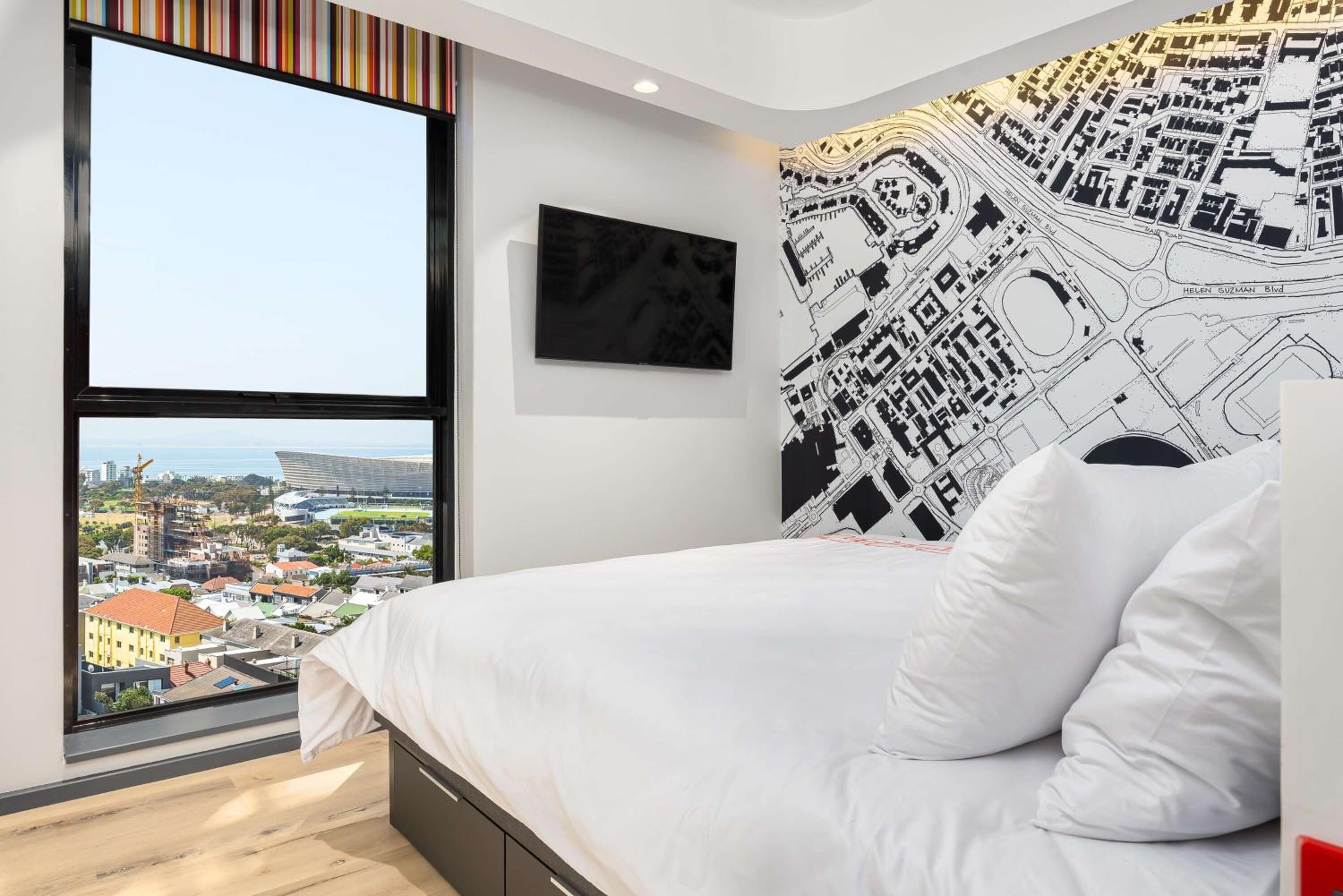 A modern bedroom with a large window, a bed with white bedding, a black and white cityscape wall art, and a framed picture on the wall.