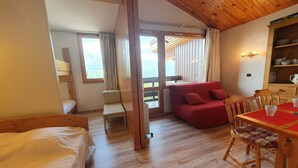 A cozy room with a wooden ceiling, a red sofa, a wooden dining table with chairs, and a view through a glass door.