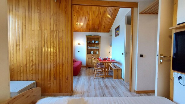 A room with a wooden floor, a bed, a dining area with a table and chairs, and a kitchen area with shelves and cabinets.