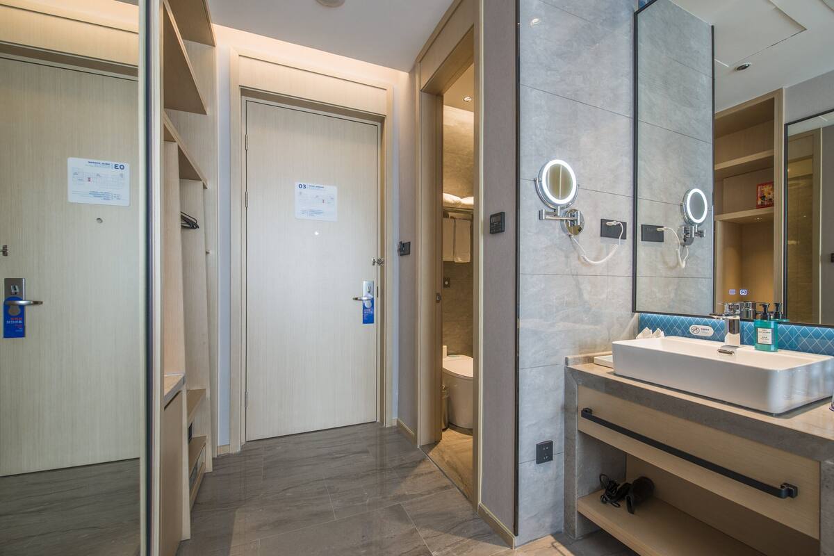 A modern hotel bathroom with a large mirror, a sink, and a toilet visible through an open door.