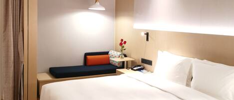 A modern hotel room with a large bed, a bedside table with a lamp, a small table with a vase of flowers, and a sofa with cushions.