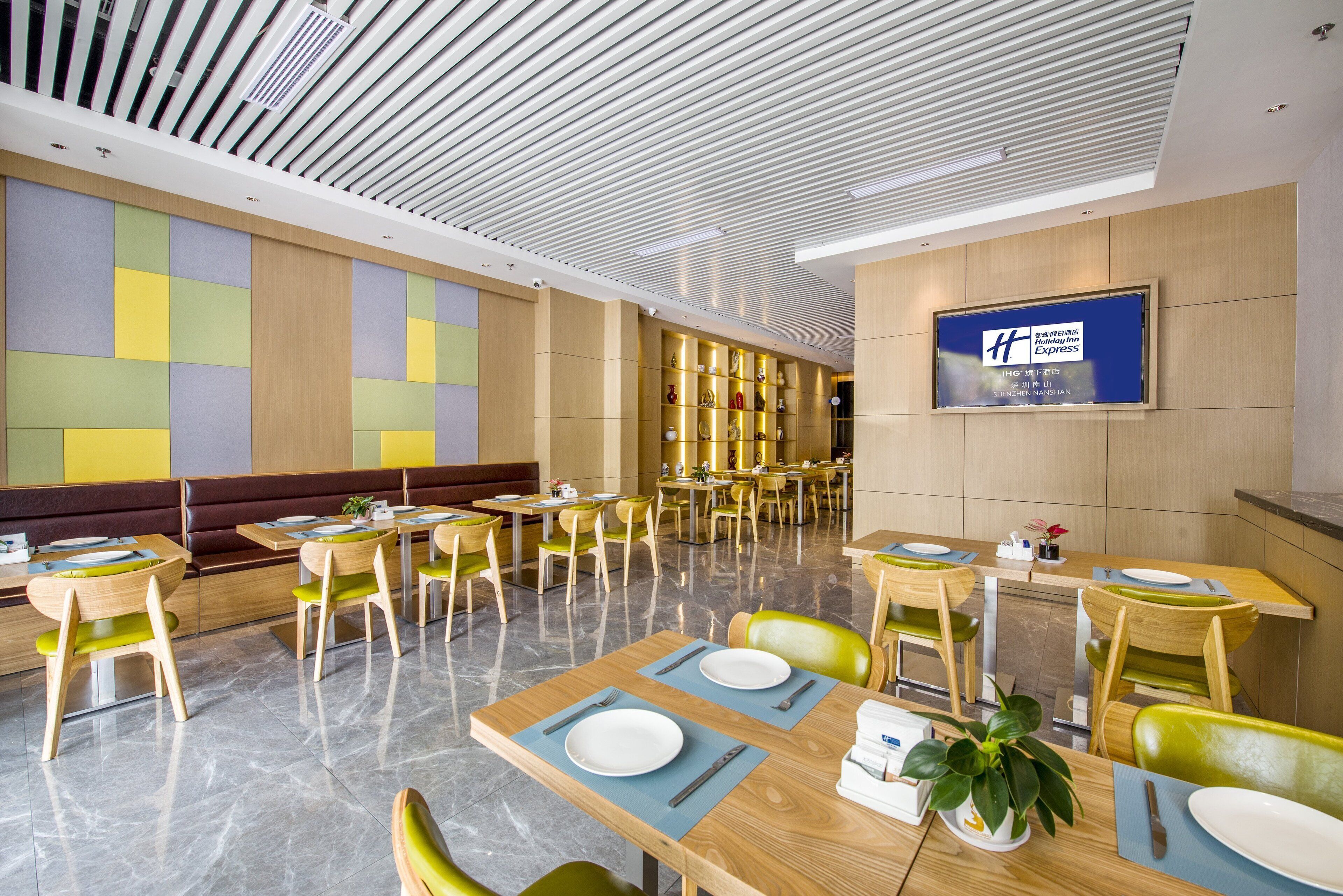 A modern dining area with wooden tables and chairs, a variety of seating options including booths and high tables, and a large screen displaying the hotel name.