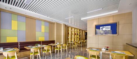 A modern dining area with wooden tables and chairs, a variety of seating options including booths and high tables, and a large screen displaying the hotel name.