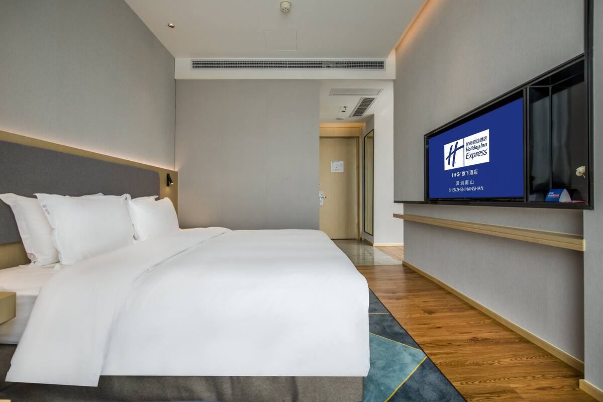A hotel room with a large bed, a flat-screen TV mounted on the wall, and a wooden floor.