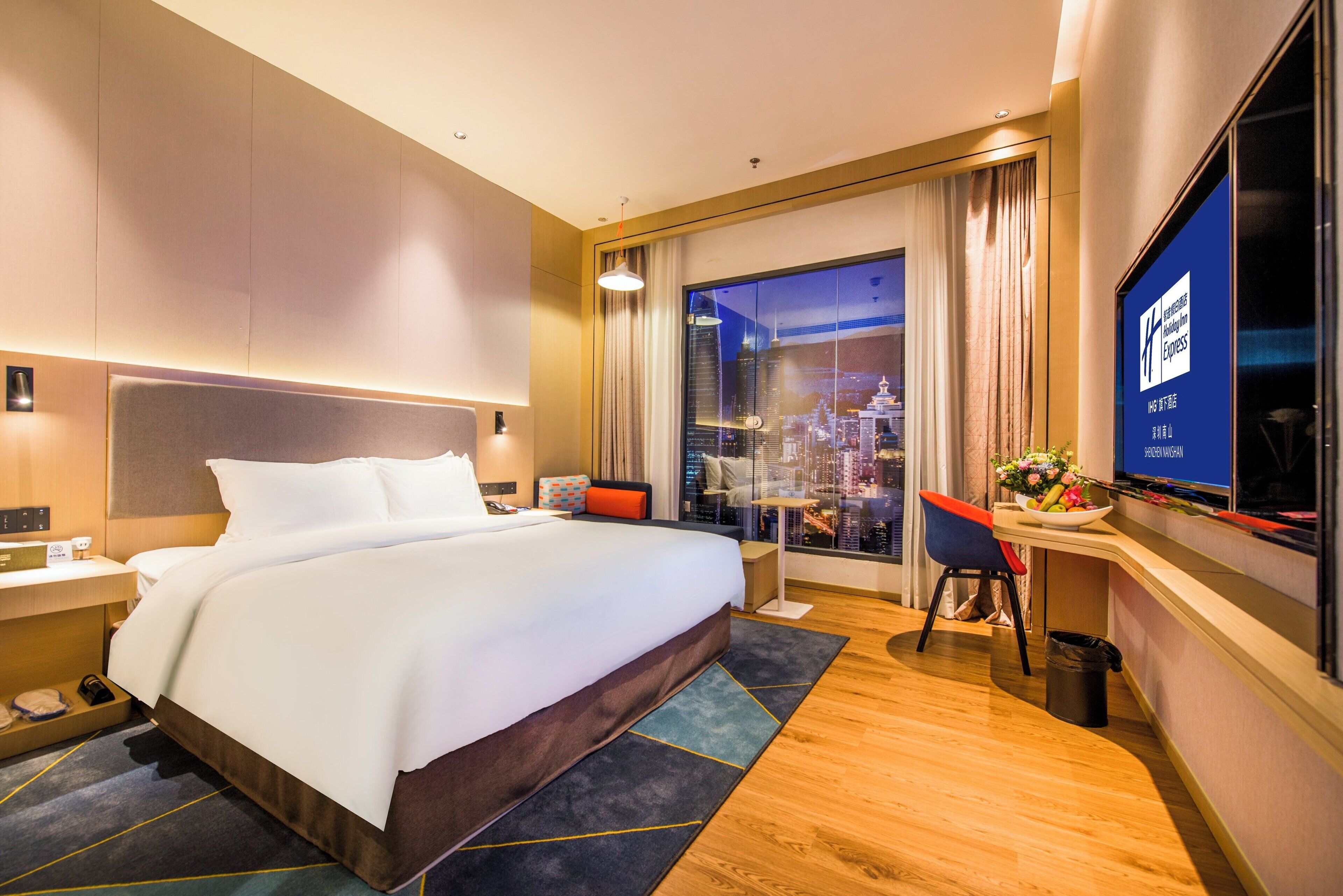 A hotel room with a large bed, a desk with a chair, a television, and a view of a cityscape through the window.