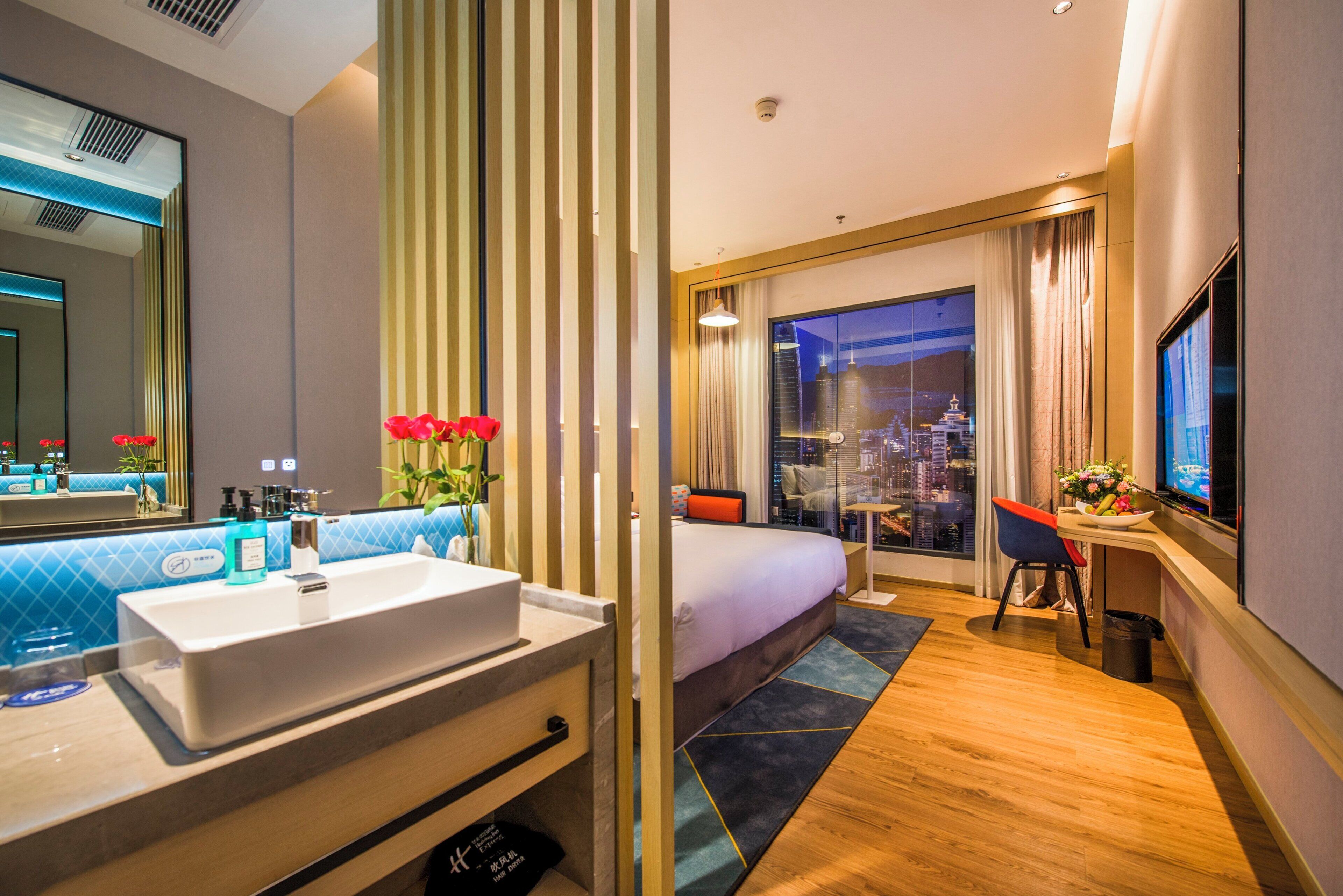 A hotel room with a large bed, a desk with a chair, a television, a bathroom with a sink and mirror, and a city view through the window.
