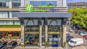 A Holiday Inn Express hotel with a modern glass facade, outdoor seating area, and a parking lot with various vehicles. - Holiday Inn Express Shenzhen Nanshan by IHG (Shenzhen)