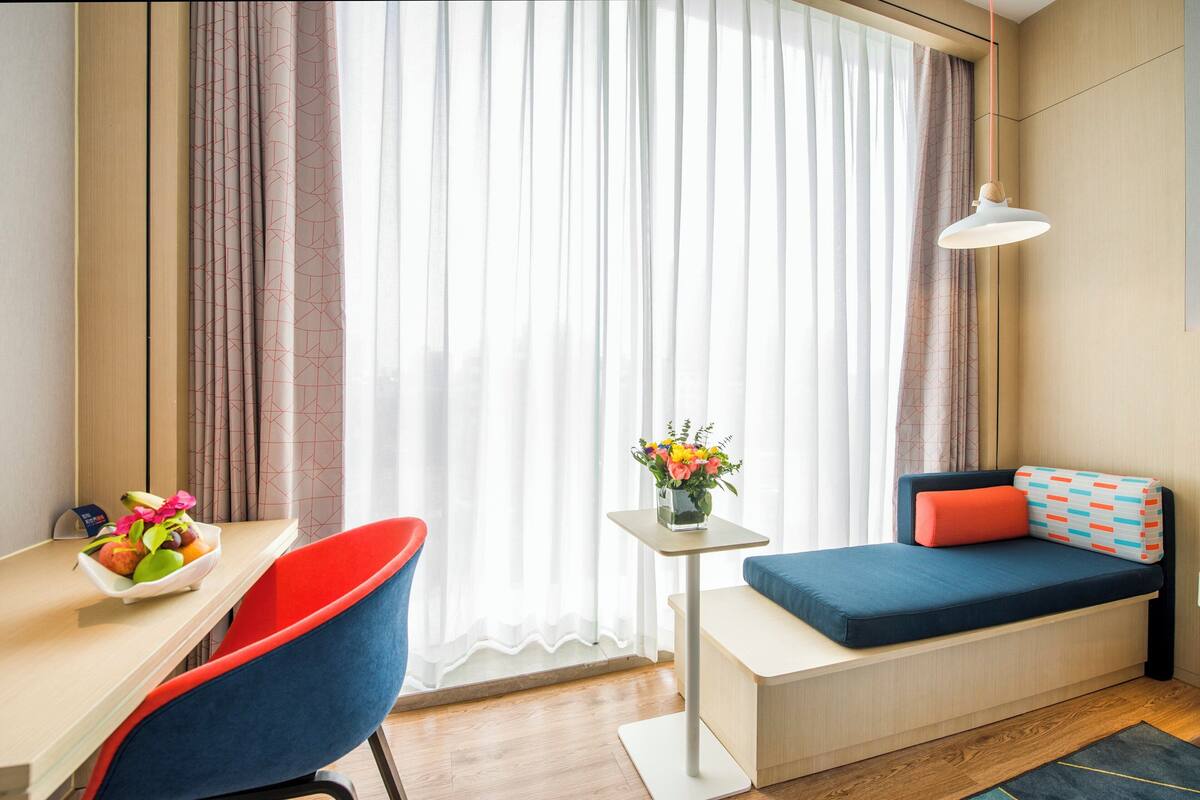 A compact room with a desk, a chair, a small table with a vase of flowers, a sofa with cushions, and a hanging light fixture.