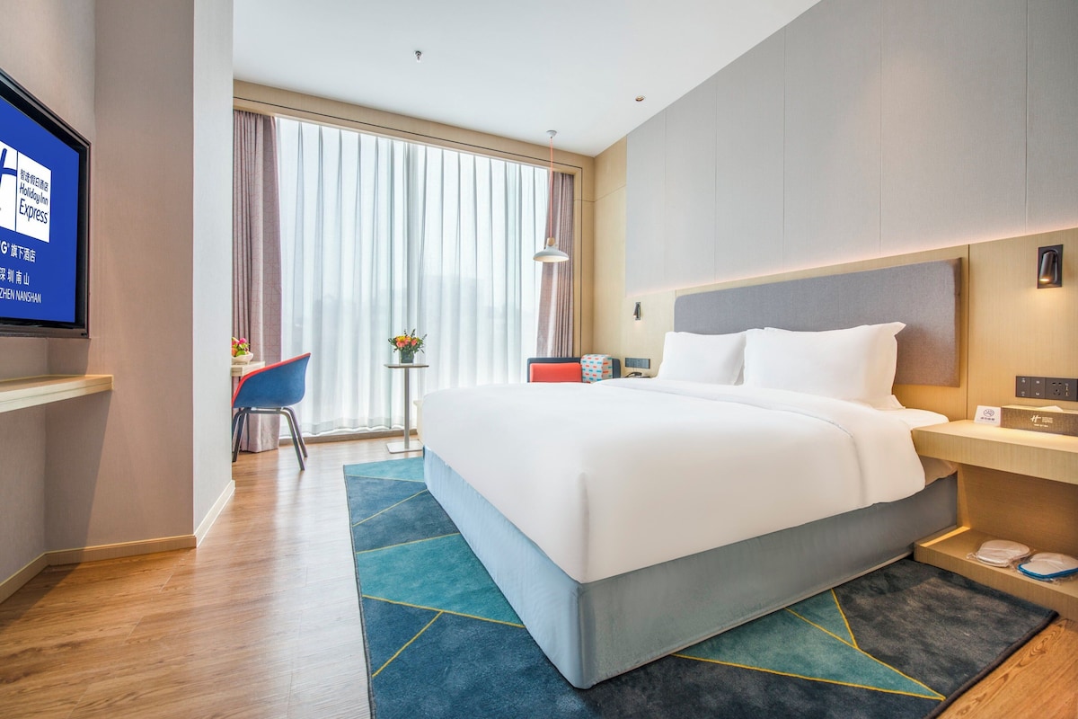 A modern hotel room with a large bed, a desk, a chair, and a TV on the wall.