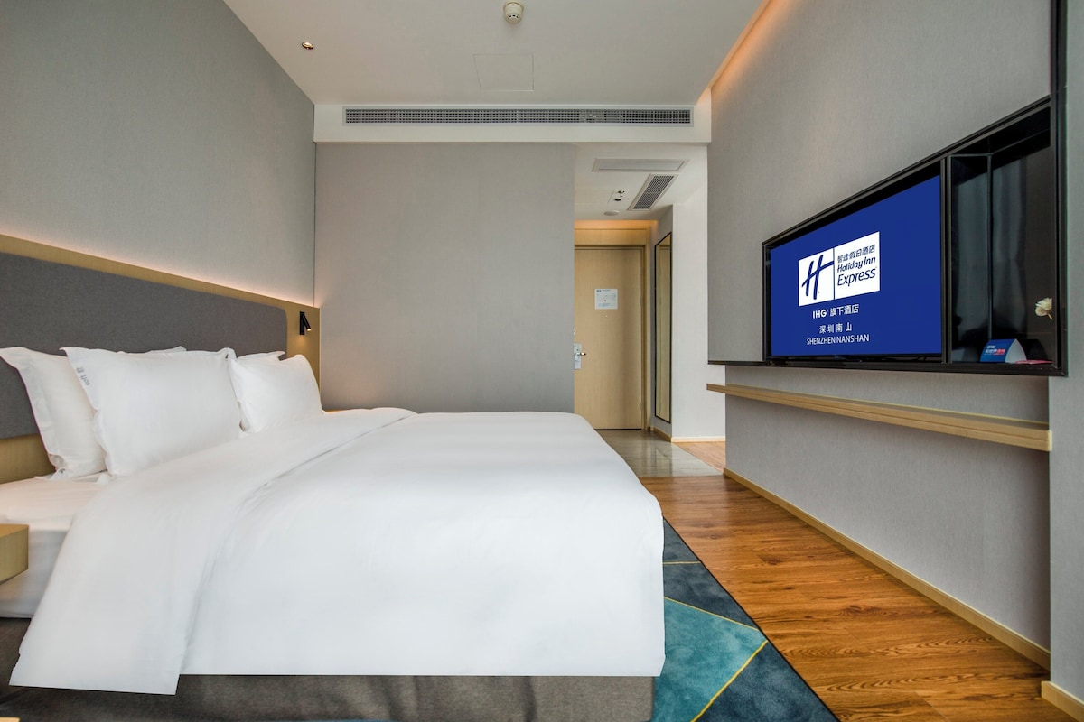 A hotel room with a large bed, a flat-screen TV mounted on the wall, and a wooden floor.