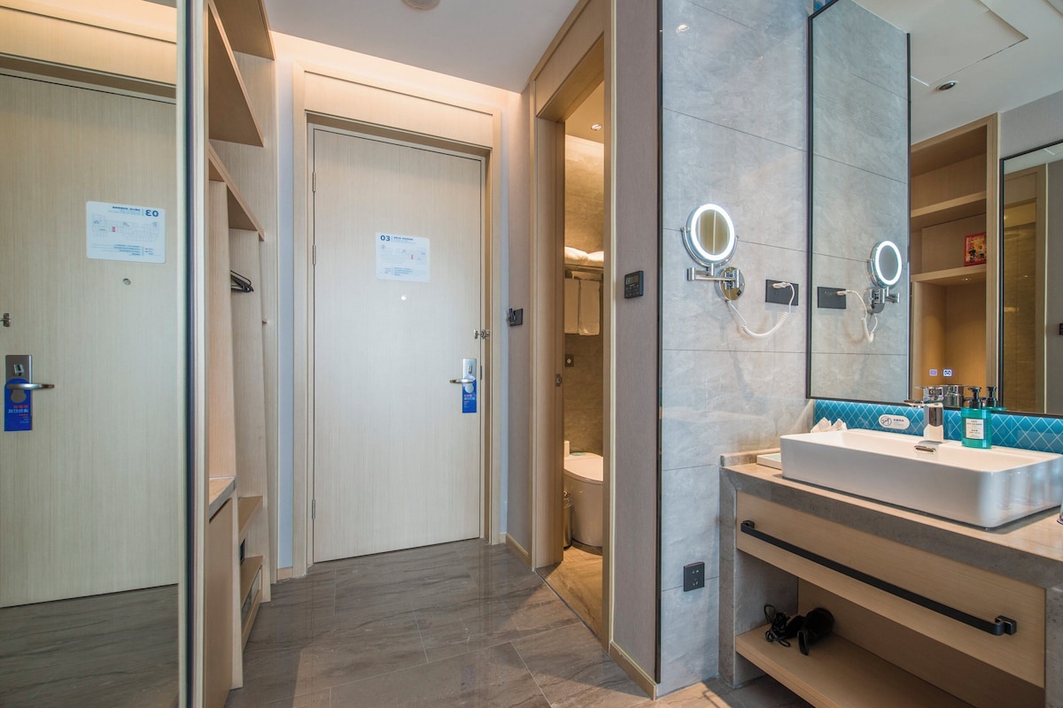 A modern hotel bathroom with a large mirror, a sink, and a toilet visible through an open door.