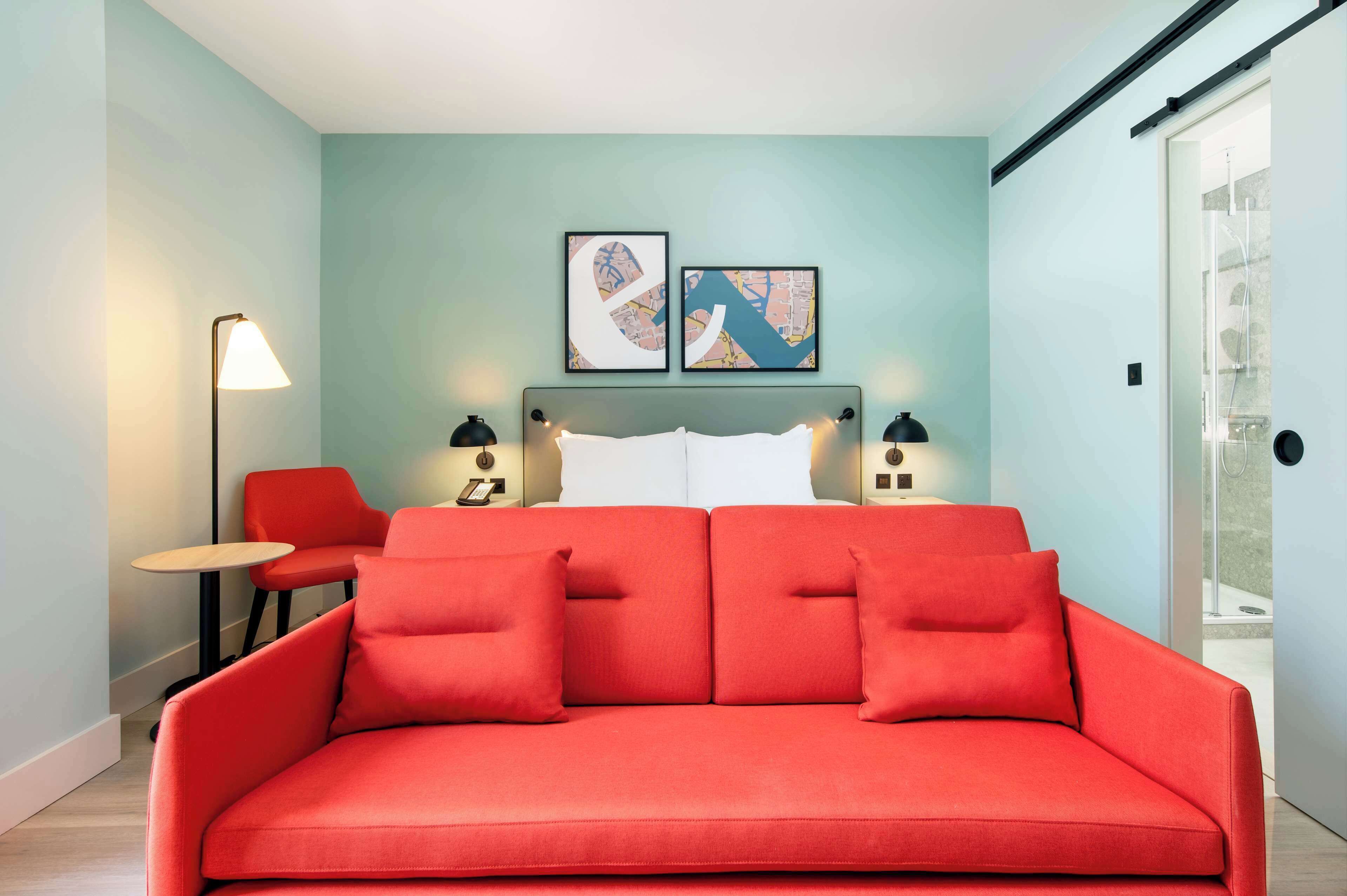 A modern room with a red sofa, a small table, and two chairs. There are two framed artworks on the wall above the sofa.