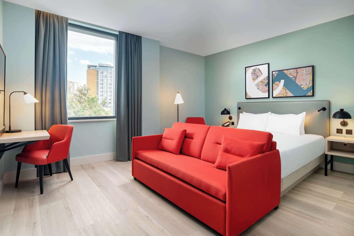 Une chambre d’hôtel avec un lit, un canapé rouge, un bureau et une chaise. Il y a deux lampes et une fenêtre avec des rideaux.