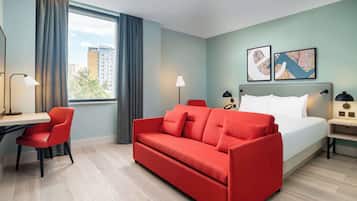 Hotellihuoneessa on sänky, punainen sohva, työpöytä ja tuoli. Huoneessa on myös kaksi lamppua ja ikkuna, jossa on verhot.