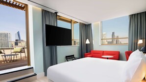 Habitación de hotel con una cama grande, un televisor en la pared, un sofá rojo, una mesita pequeña y vistas a la ciudad por la ventana.