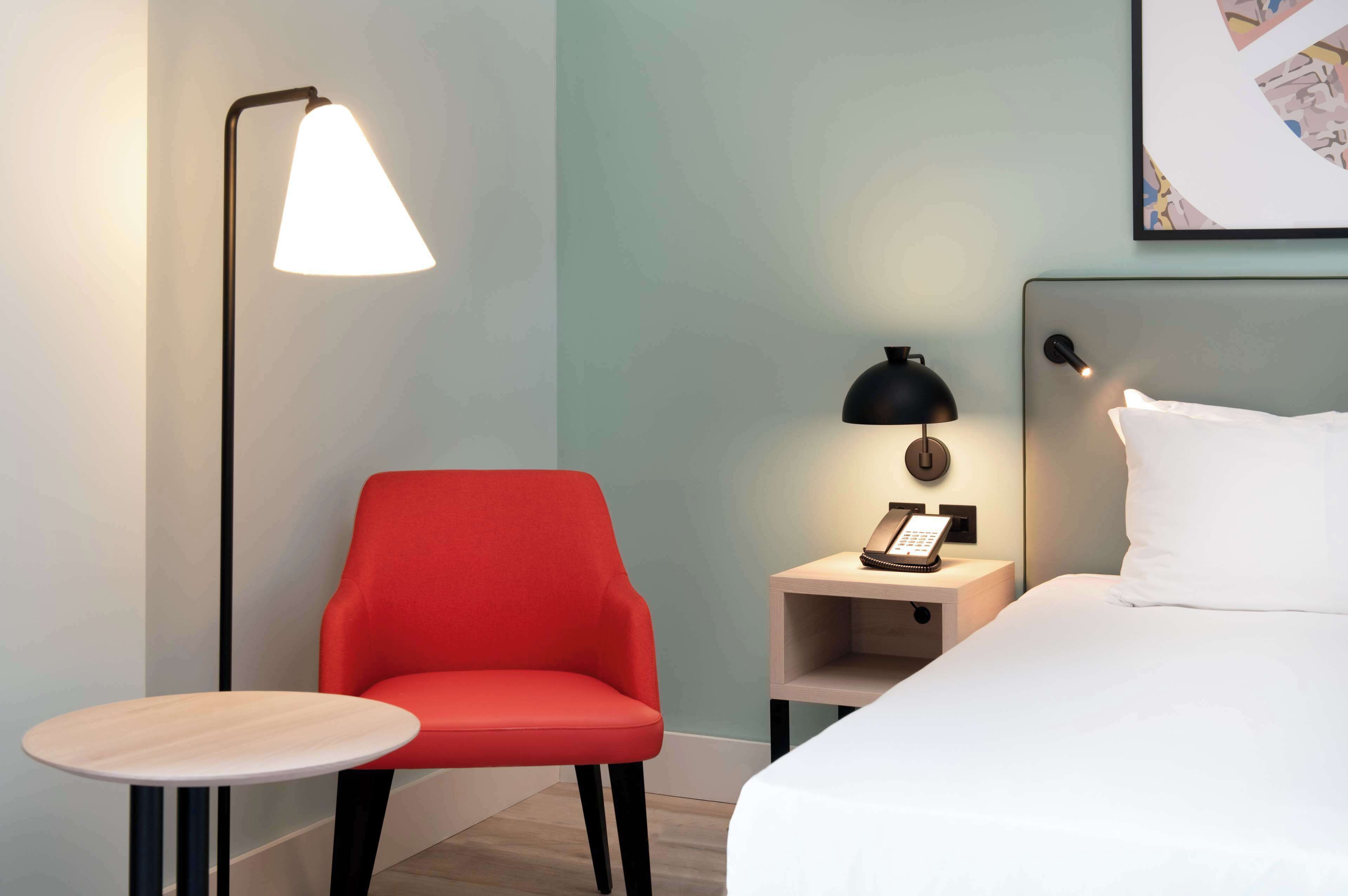 A hotel room with a bed, a red chair, a small table, a bedside lamp, and a nightstand with a phone.