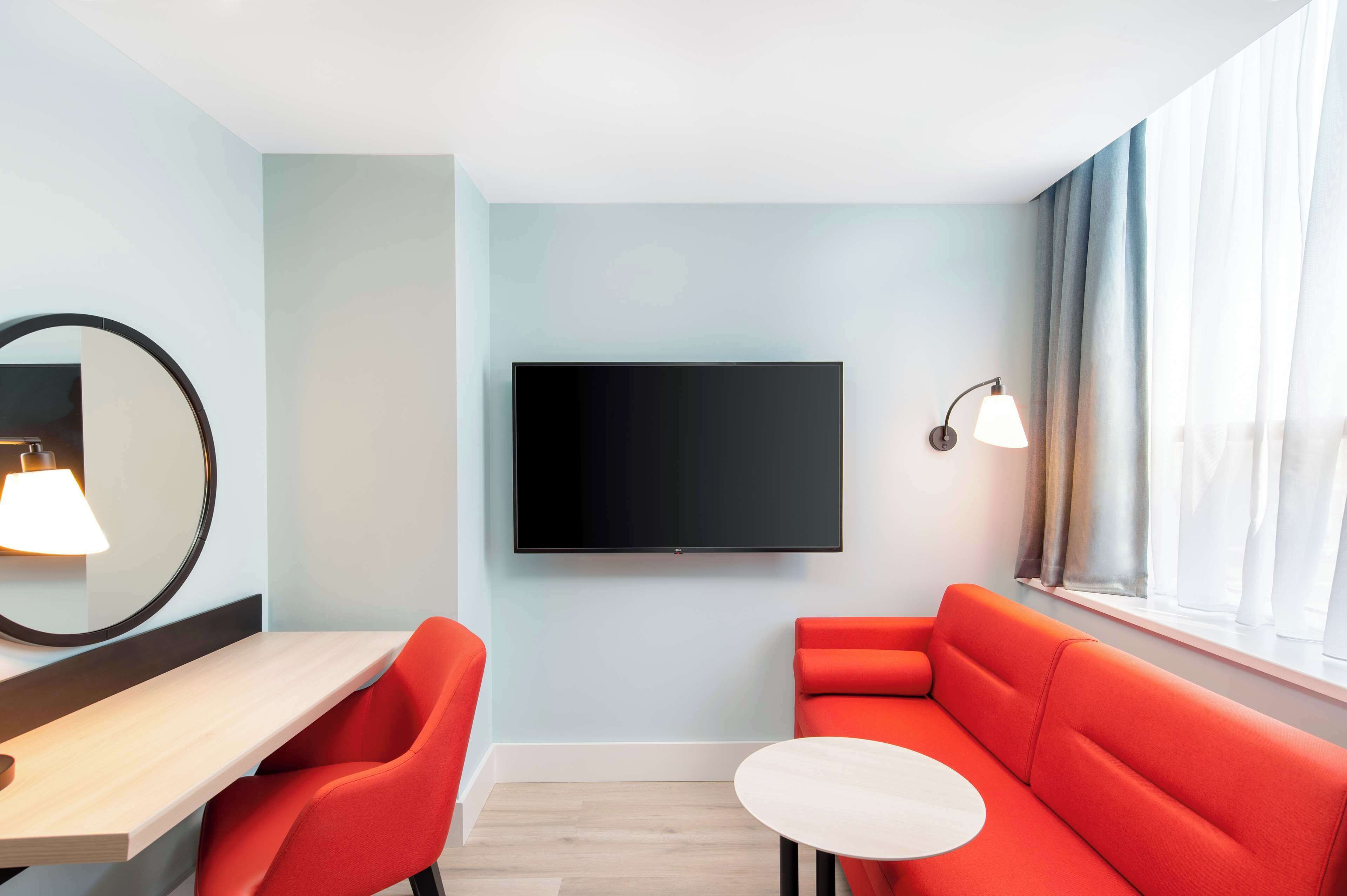 Ein modernes Zimmer mit einem wandmontierten Flachbildfernseher, einem Holztisch, zwei roten Stühlen, einem kleinen weißen Tisch und einem fenster mit Vorhängen.