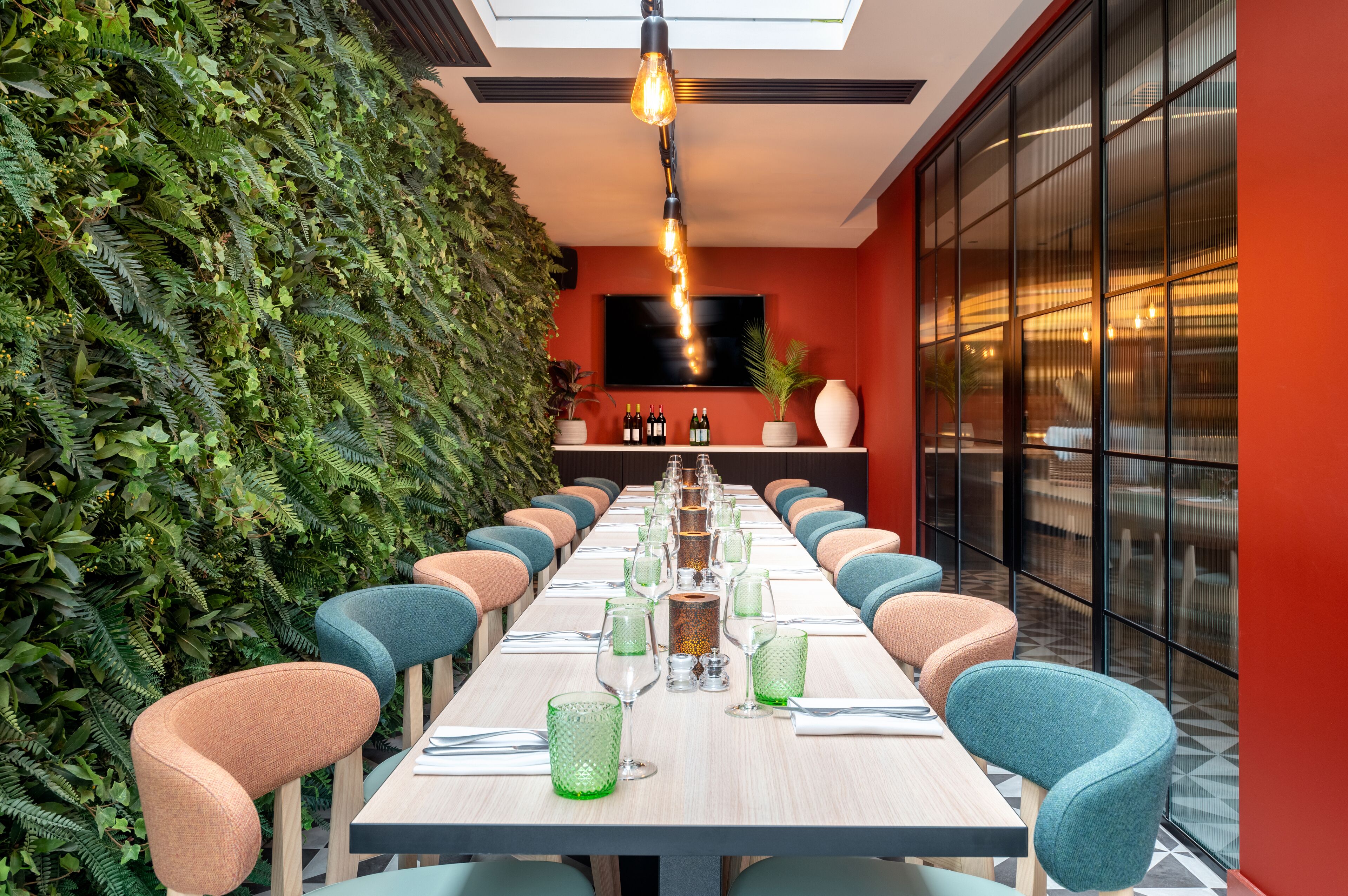 A long dining table with chairs, a TV mounted on the wall, and a green wall with plants.