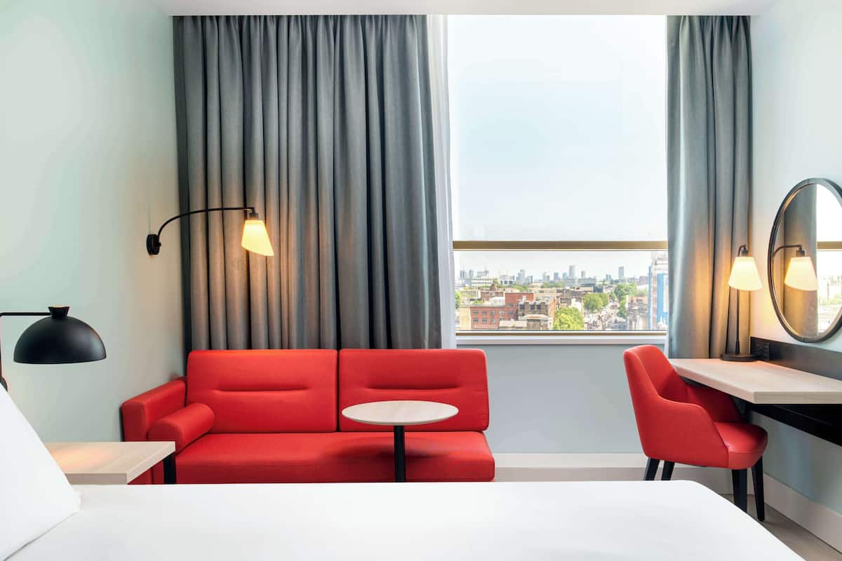 Une chambre d’hôtel avec un canapé rouge, une petite table ronde, un bureau avec une chaise, un lit et une vue sur la ville à travers de grandes fenêtres ornées de rideaux gris.