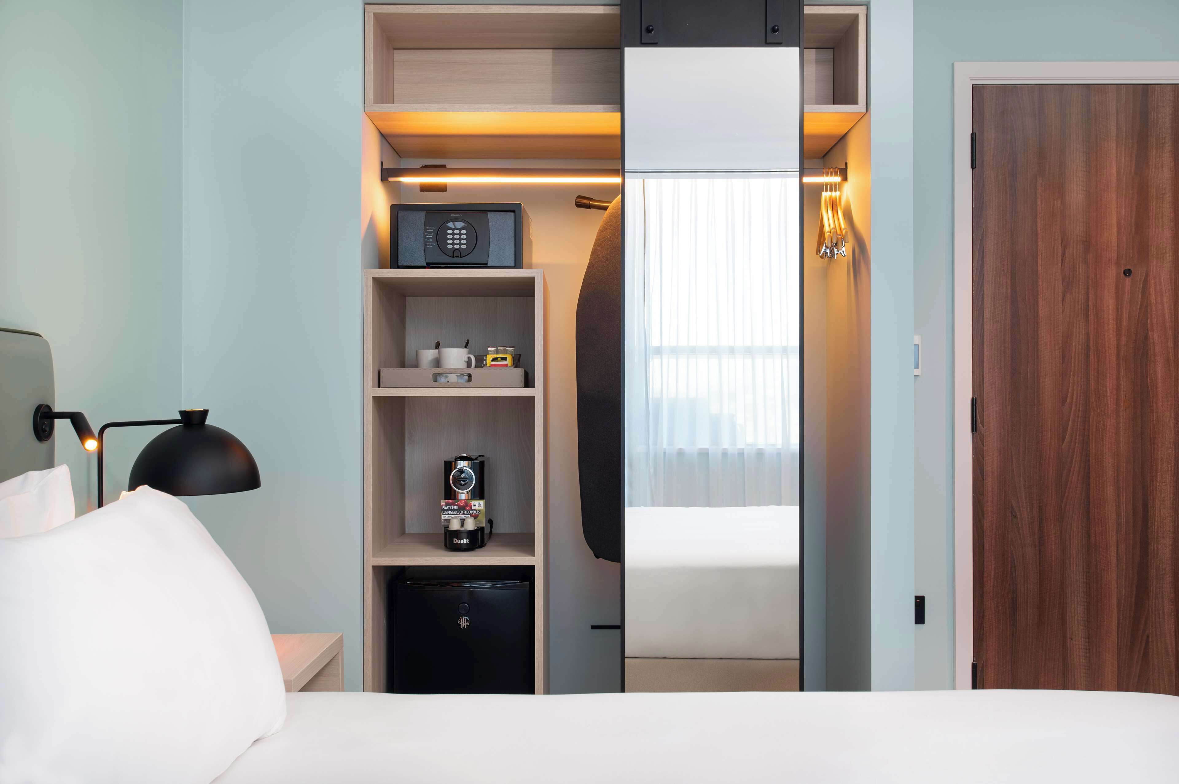 Habitació d'hotel amb llit, escriptori amb làmpada, nevera minibar, cafetera i armari.