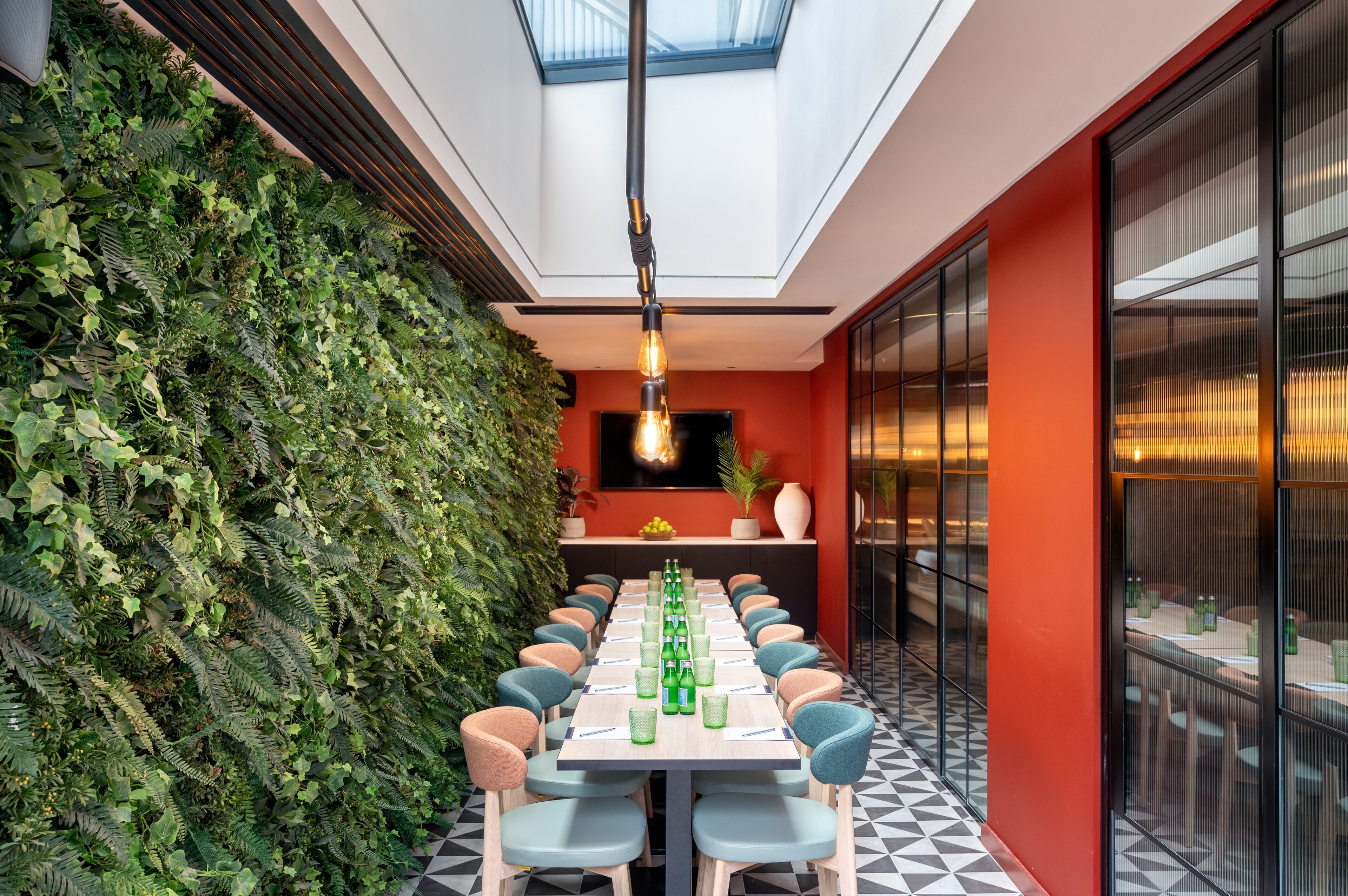A long table with chairs, a green wall with plants, and a ceiling light.