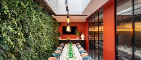 A long table with chairs, a green wall with plants, and a ceiling light.