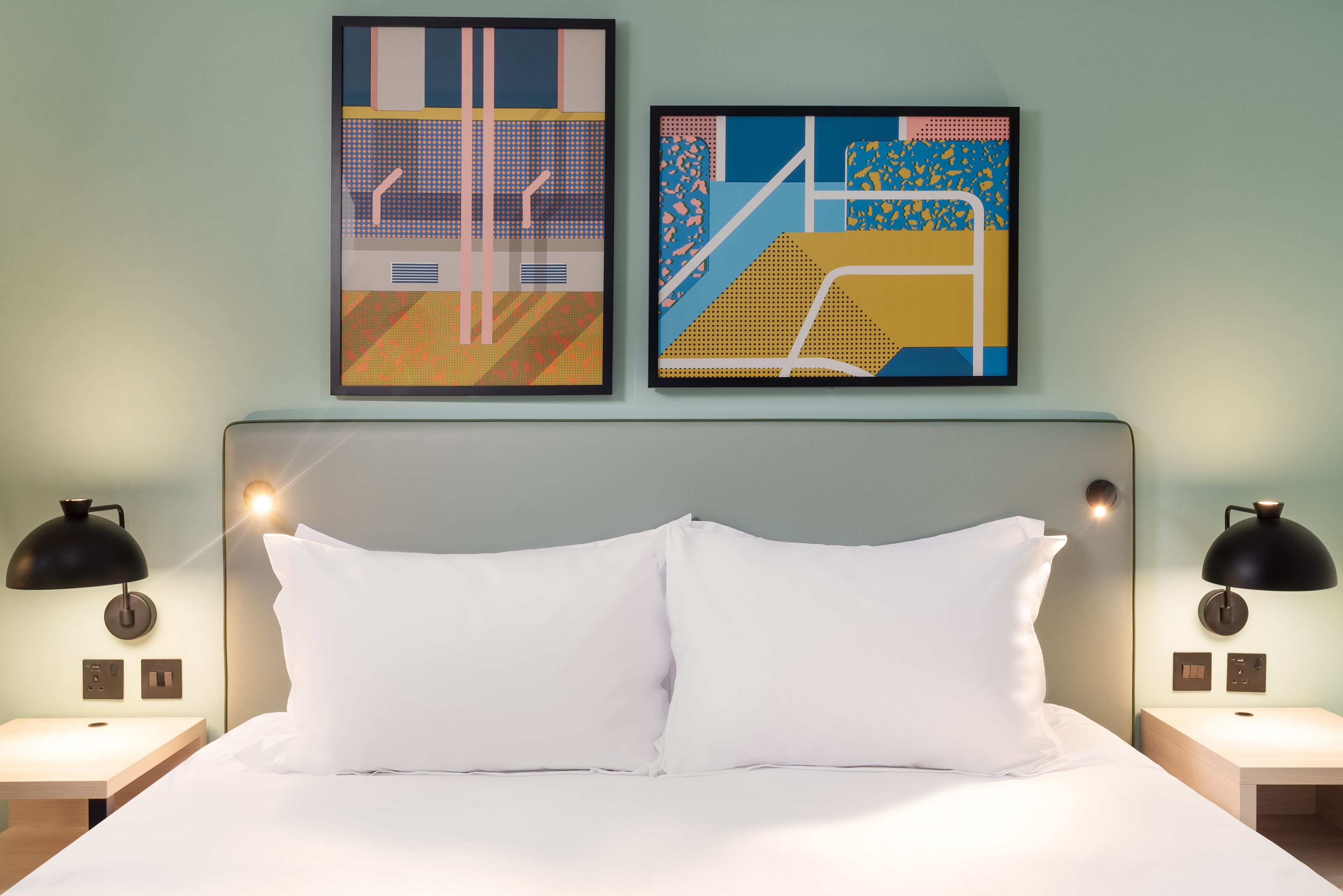 A neatly made bed with white bedding, two black wall-mounted lamps, and two abstract paintings above the bed.