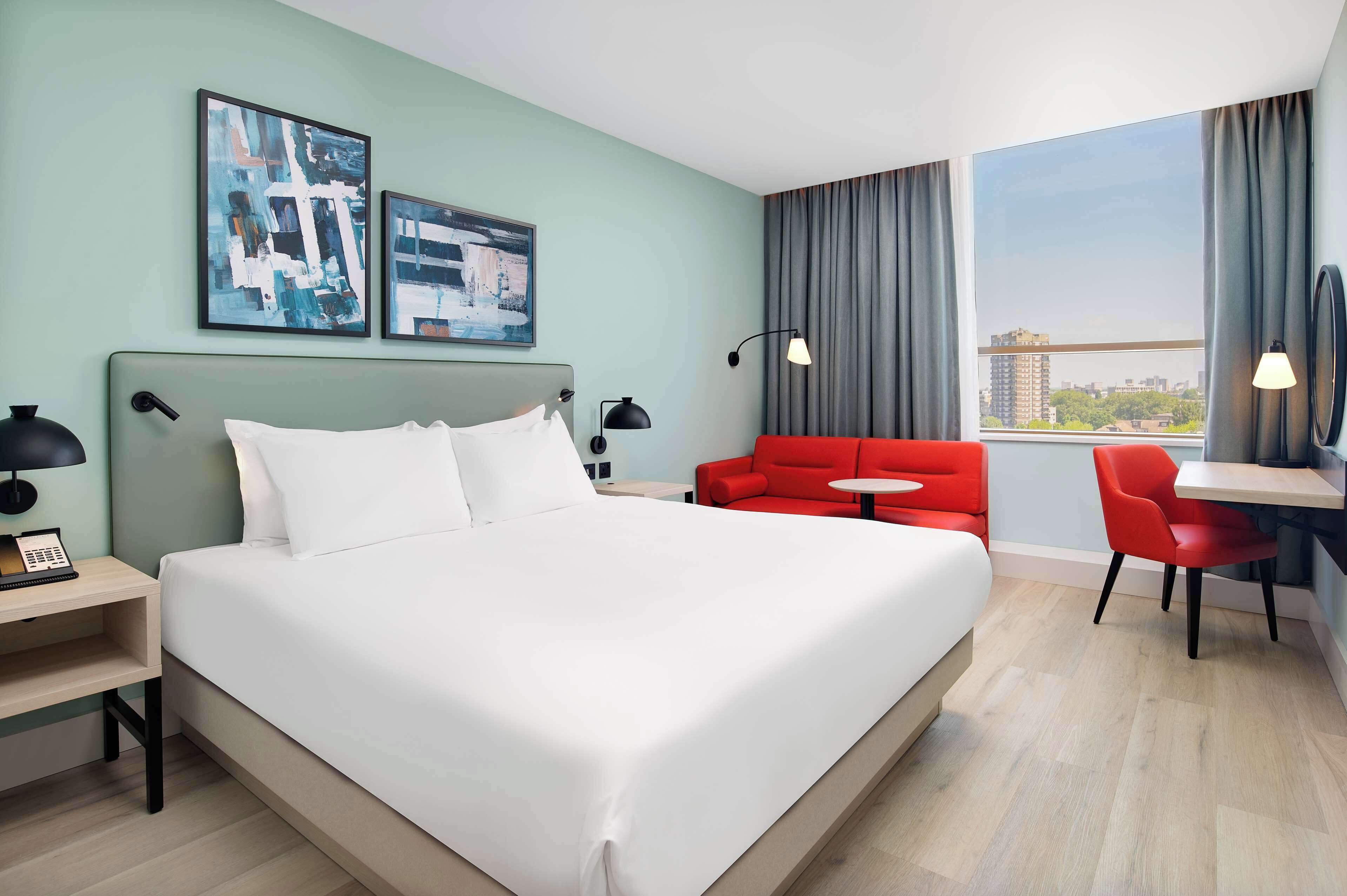 Een hotelkamer met een groot bed, een rode bank, een klein tafeltje en uitzicht op de stad door het raam.