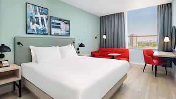 A hotel room with a large bed, a red sofa, a small table, and a view of the city through the window.