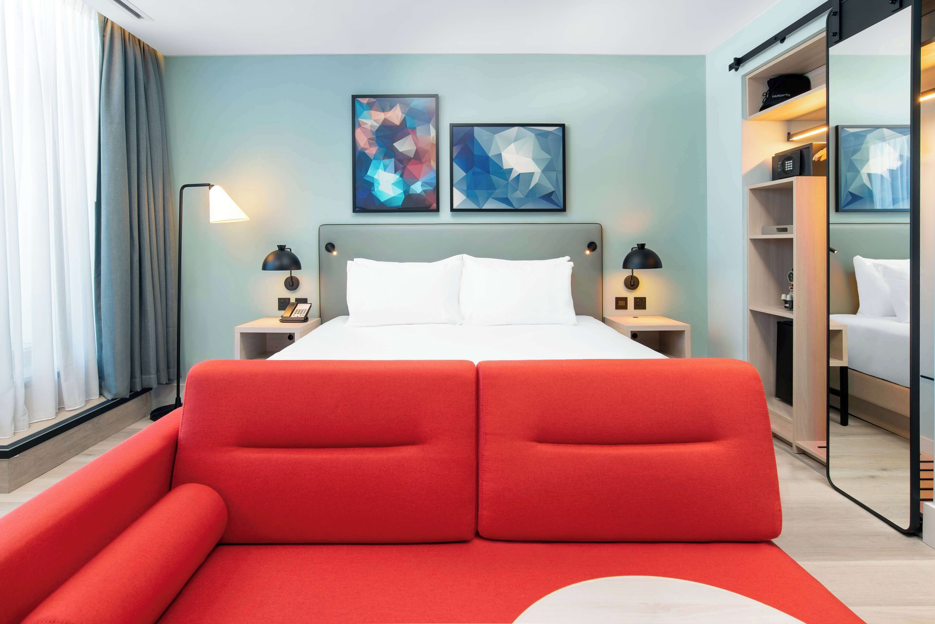 A modern hotel room with a large bed, two bedside tables, a red sofa, and artwork on the wall.