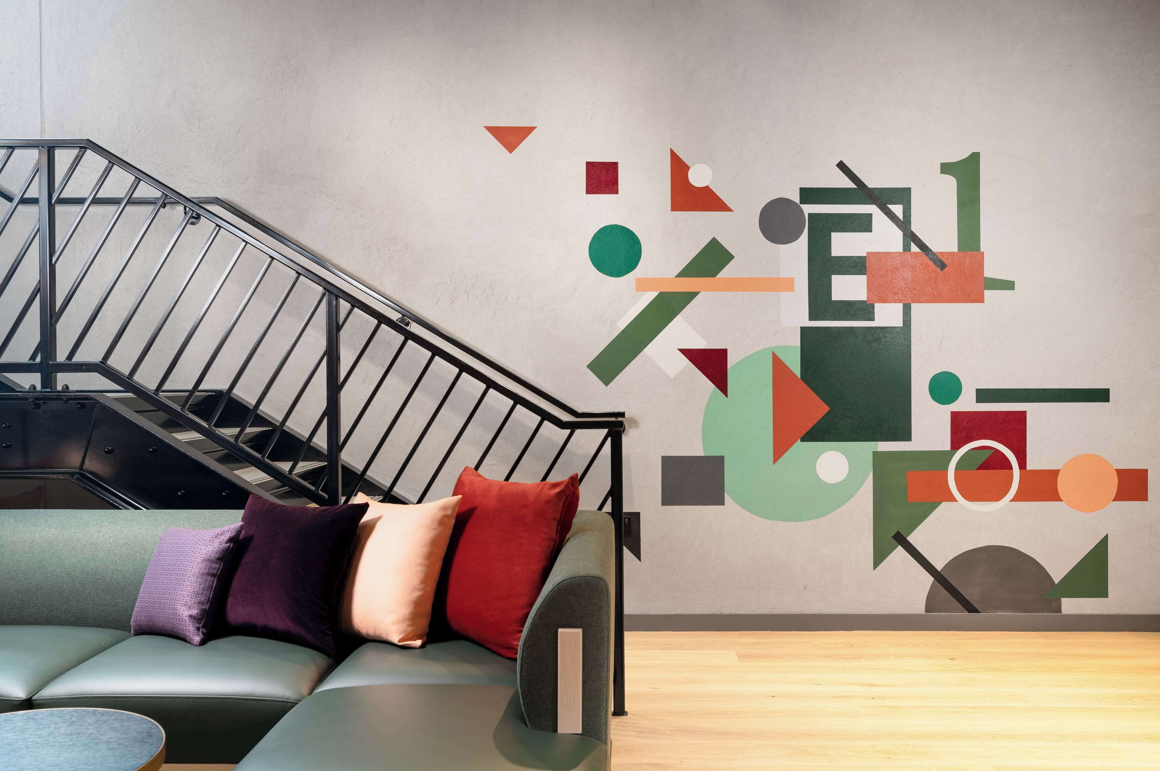 A modern interior with a staircase, a green sofa with cushions, and a geometric wall mural.