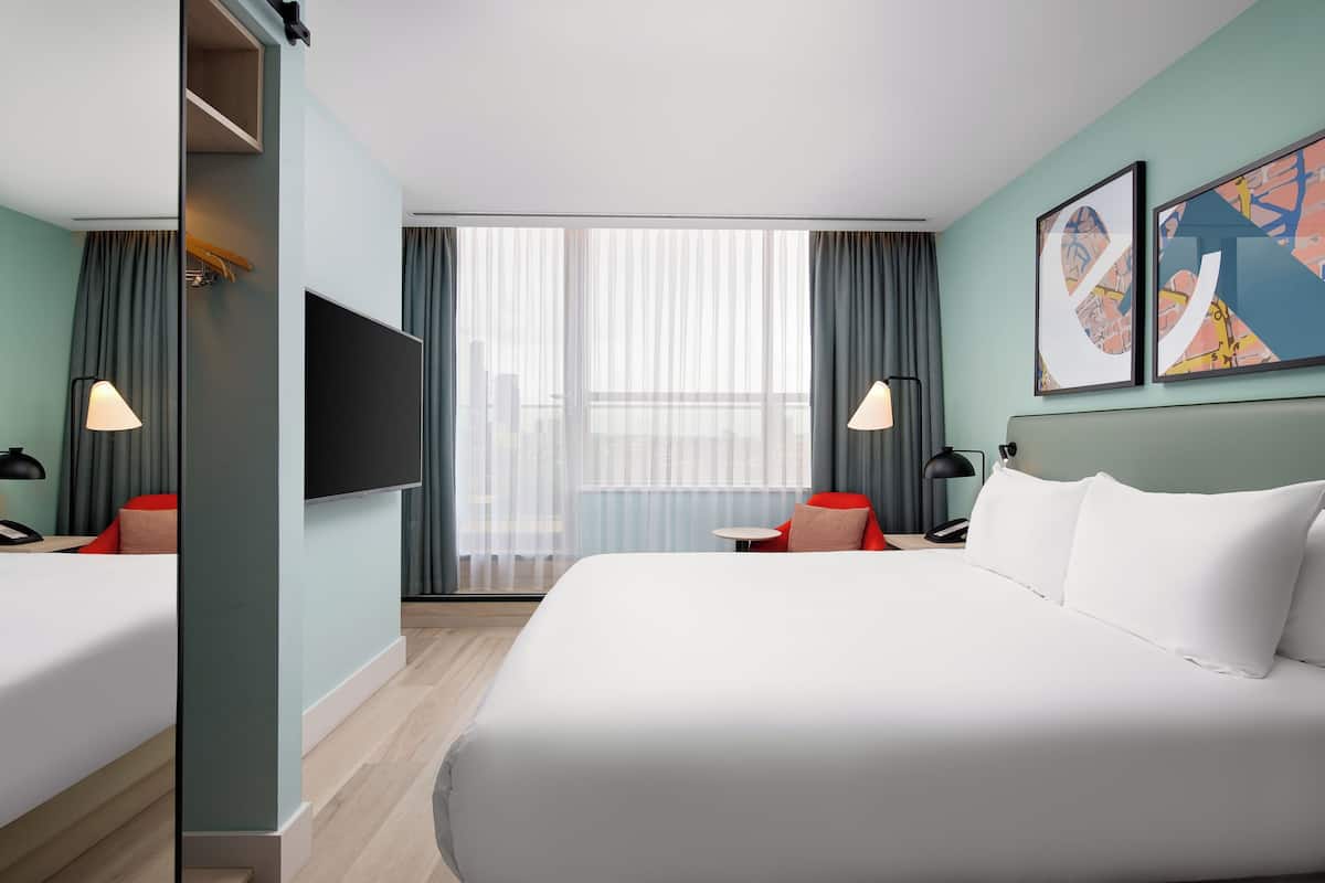 Une chambre d’hôtel dotée d’un grand lit, de deux tables de chevet, d’un téléviseur à écran plat et offrant une vue sur la ville grâce à de grandes fenêtres.