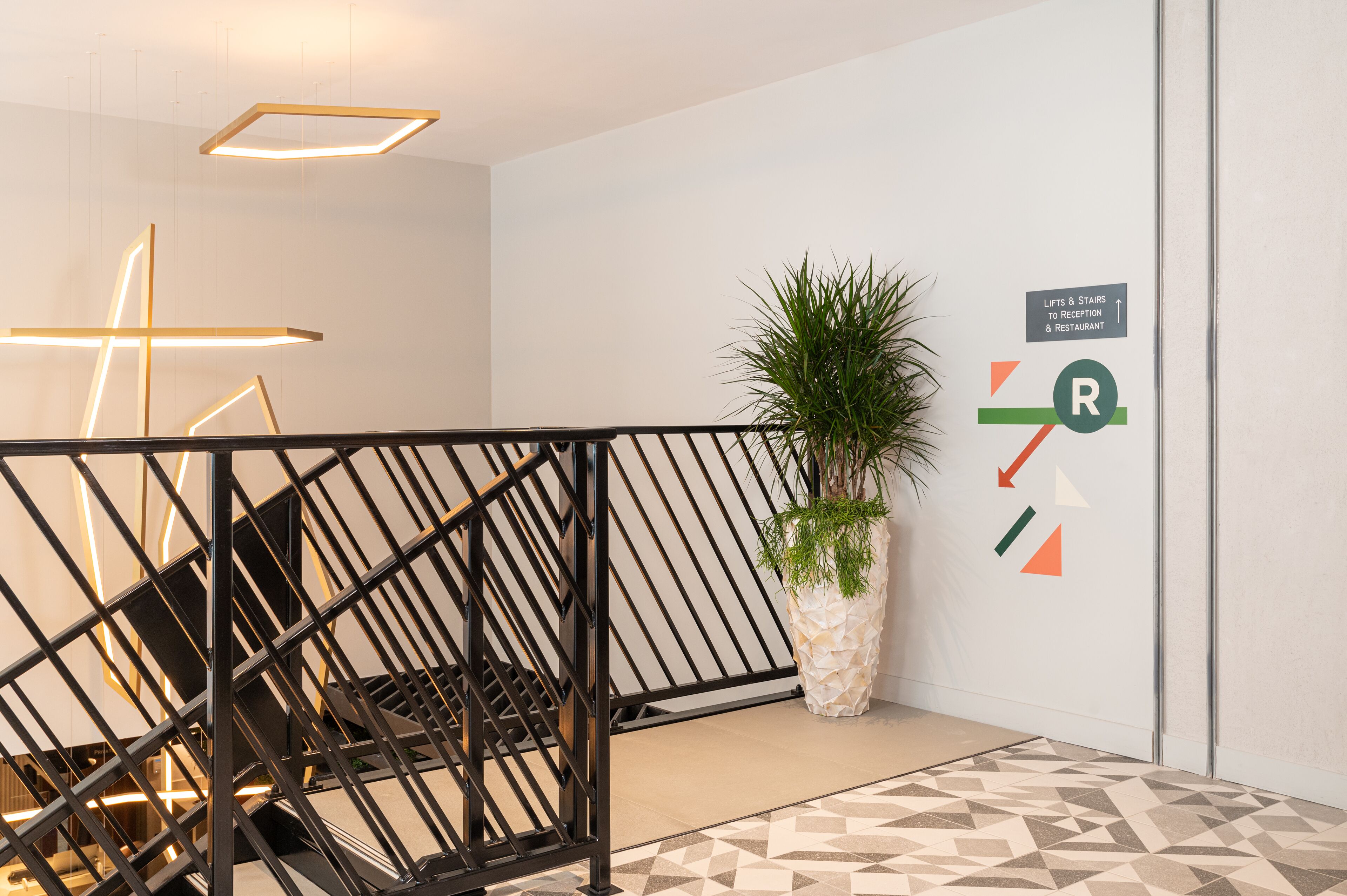 A modern interior with a geometric-patterned floor, a staircase with a black railing, and a wall-mounted sign indicating the location of the reception and restaurant.