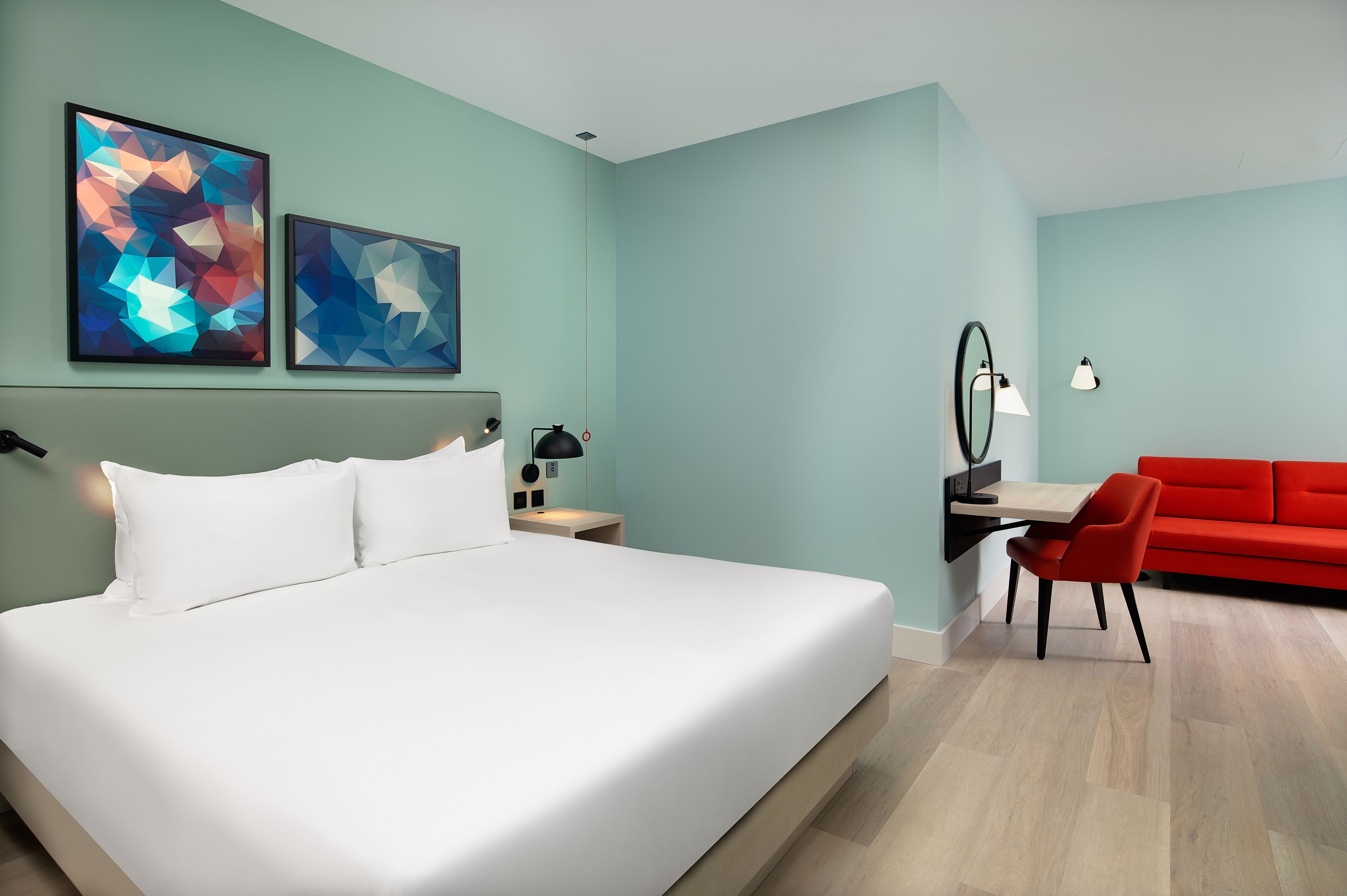 Une chambre d’hôtel moderne avec un grand lit, un bureau avec une chaise, un canapé rouge et des œuvres d’art abstraites accrochées au mur.