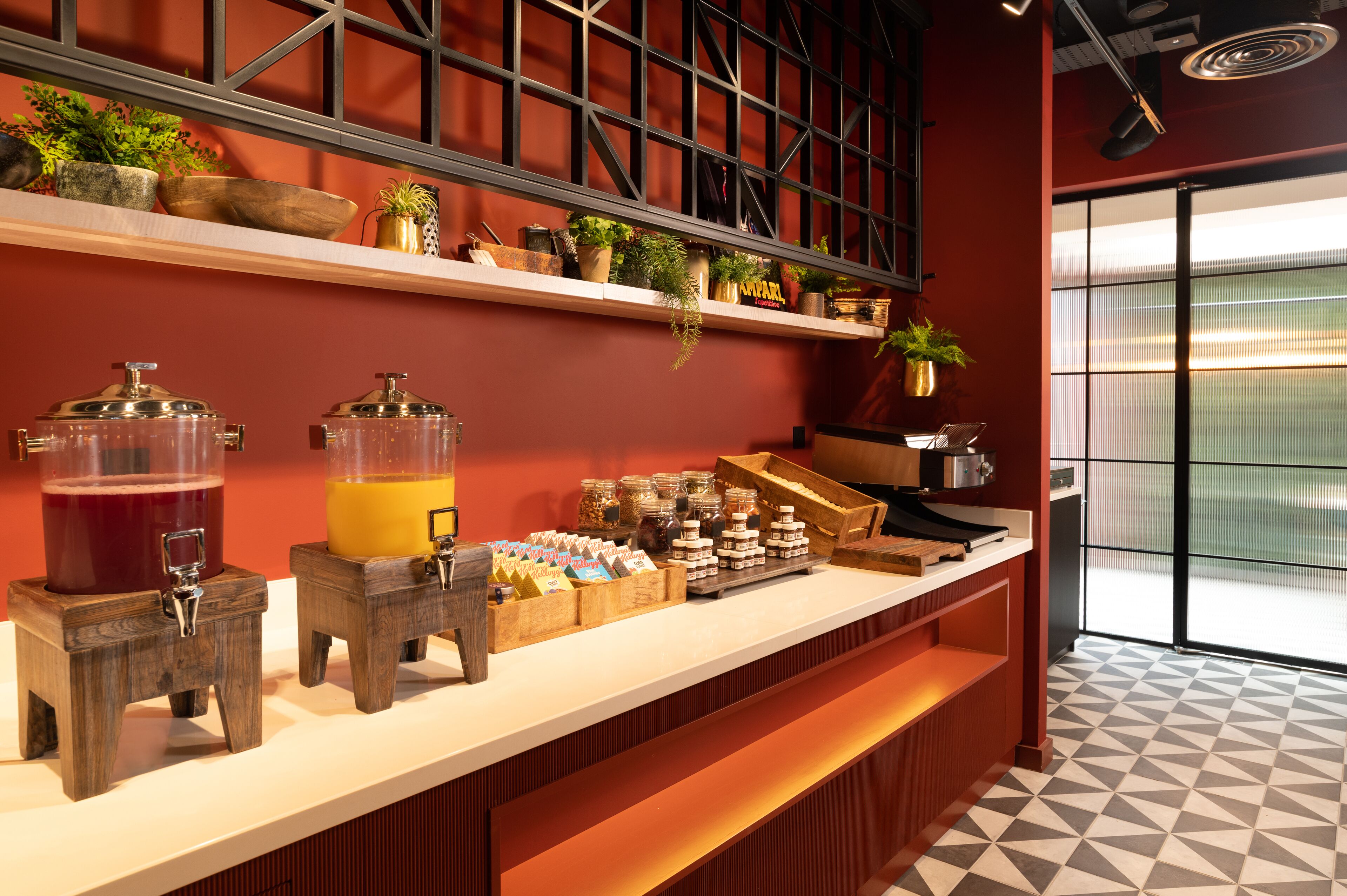 A breakfast bar with two beverage dispensers, a variety of food items, and shelves with plants.