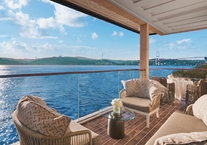 A balcony with wicker furniture, a glass coffee table, and a view of a body of water with a bridge in the distance.