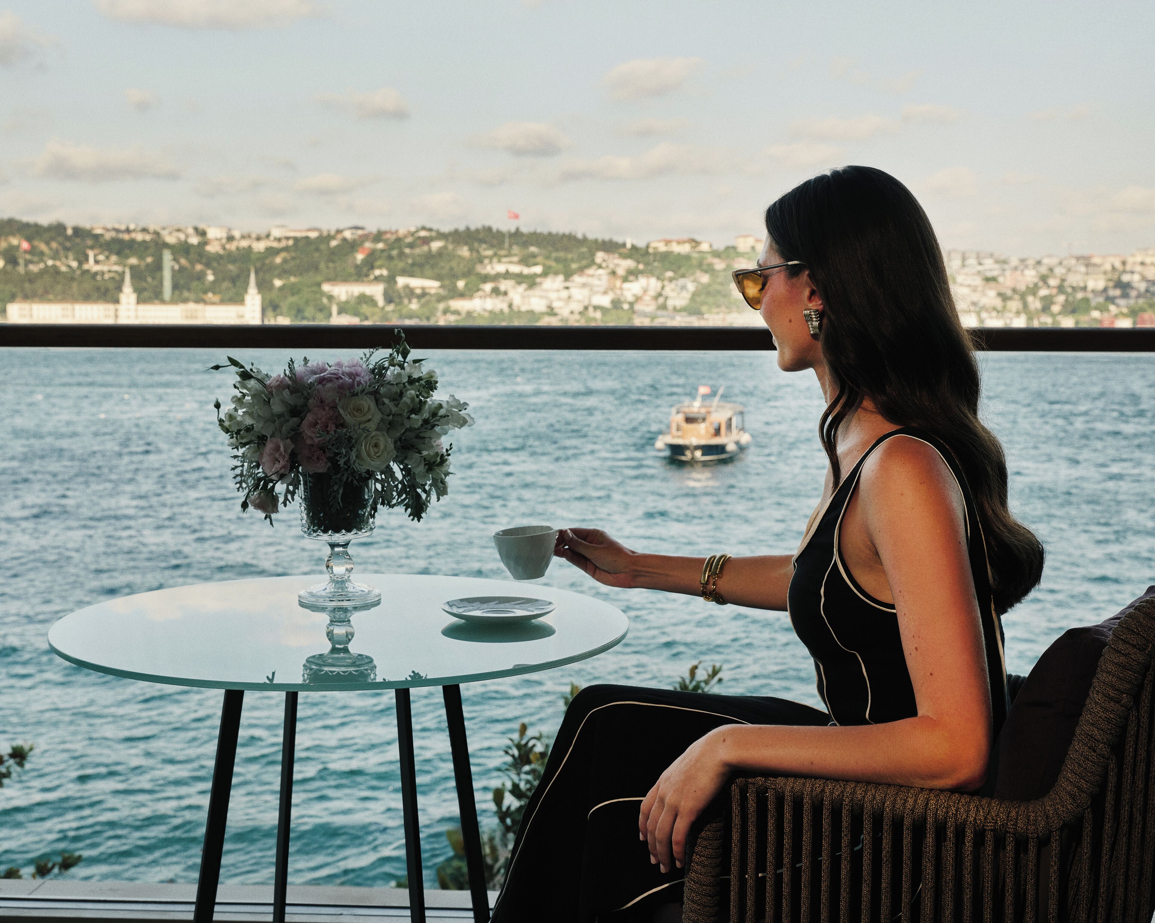 A woman seated at a table with a vase of flowers, overlooking a body of water with a boat in the distance.