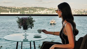 A woman seated at a table with a vase of flowers, overlooking a body of water with a boat in the distance.